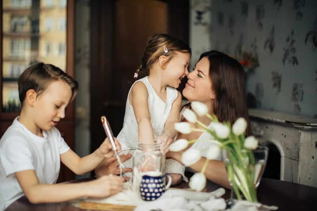 Smiling mom and daughter rub noses while son plays next to them.