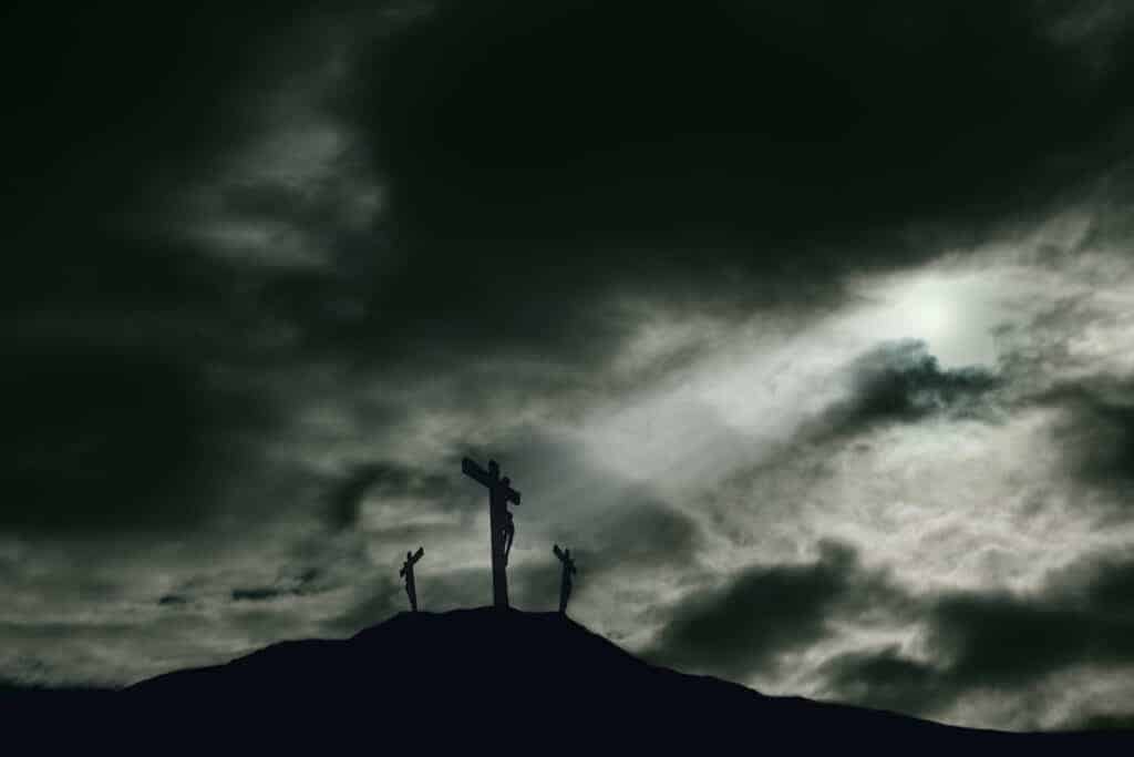 A depiction of the crucifixion of Jesus Christ on a cross with 2 other robbers nearby on Calvary. The sky is darkened and rays of light break through the clouds onto the cross for drama. Concept of the death of Jesus on Good Friday and His resurrection on Easter Sunday. Horizontal orientation with copy space.