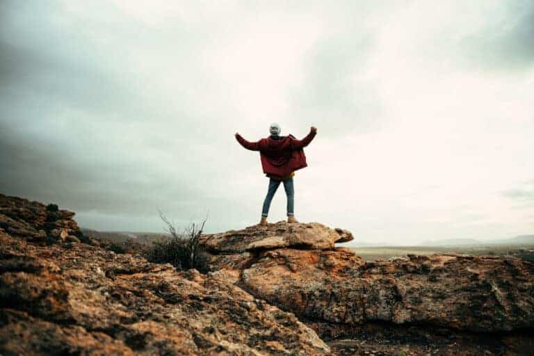 Male hiker reaching top of mountain peak with open arms embracing arms.