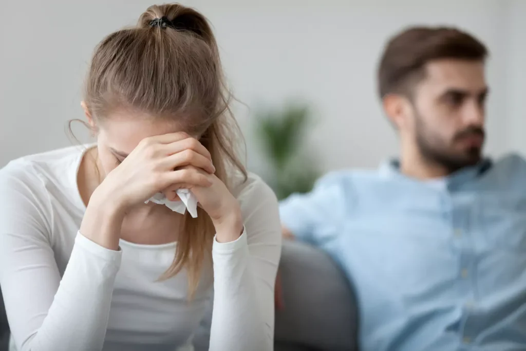 A man and a woman sit next to each other on a couch, with the man looking away off in the distance while the woman lowers her head and wipes away tears.