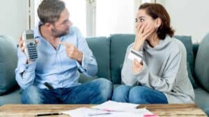 A husband and wife having a difficult discussion while sitting on the couch. The man holds up a calculator, pointing at it in frustration. The woman covers her mouth with her hand. He is having trouble forgiving his spouse for her reckless spending.