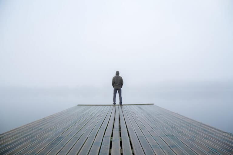 Young man standing alone on edge of footbridge and staring at lake. Mist over water. Foggy air. Early chilly morning in autumn. Beautiful freedom moment and peaceful atmosphere in nature. Back view.