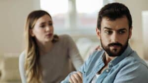 A woman, out of focus, apologizing to her spouse. Her hand is on his shoulder, but he is turned away from her, in focus, and looks like he doesn't know how to forgive her.