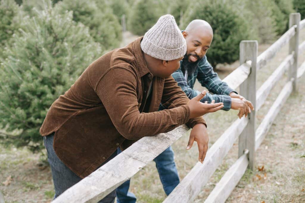 Dad and teen son standing at fence. Son is talking, dad is listening and smiling.