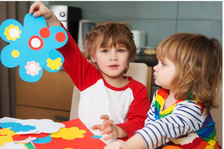 Two homeschooling children doing crafts. One is holding up a paper blue flower.