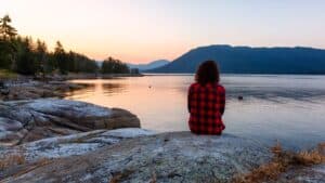 photo of a woman in plaid sitting peacefully by the water at sunset. Her spiritual disciplines have led her here and have positively impacted her mental health.