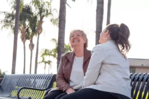 Mother and daughter laughing on park bench under palm trees.