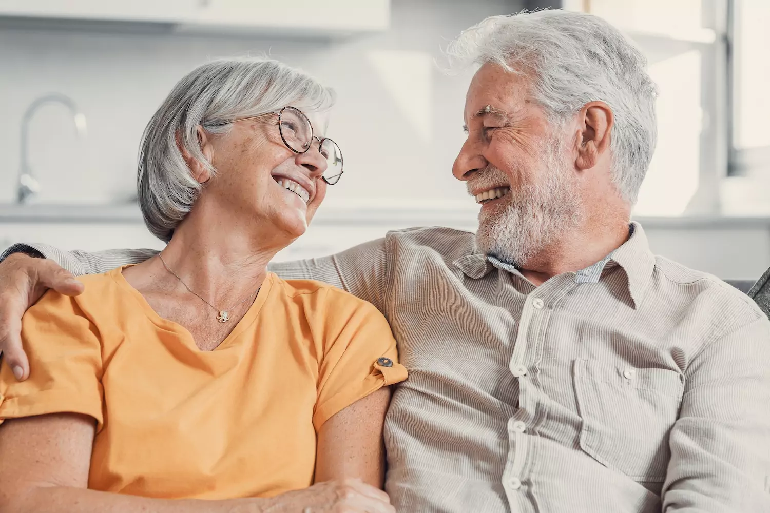 A mature, empty nest couple sit on a couch and look adoringly into each other's eyes.