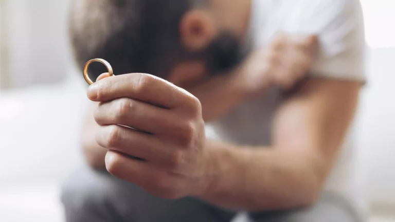 A man has removed his wedding ring, holding it up with one hand and covering his face with the other. His spouse was abusive and he has lost her by setting boundaries for his own safety. He wrestles with guilt over if he did the right thing.