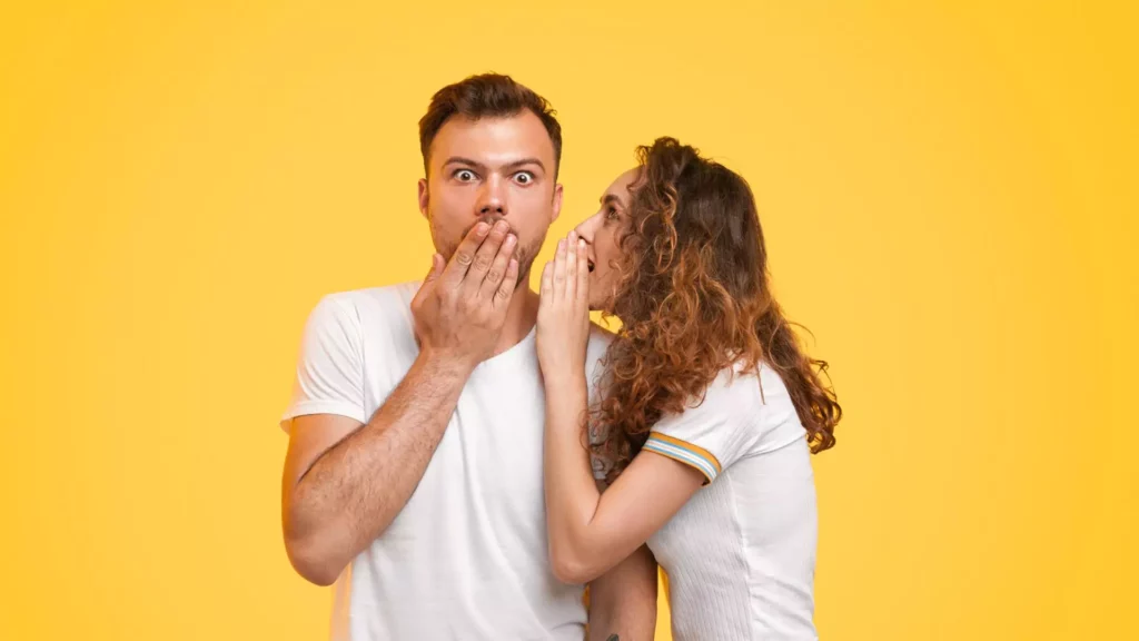 A young woman whispers into her fiancé’s ear while he looks surprised, representing the excitement and curiosity of asking important questions to ask your fiancé.