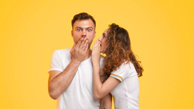 A young woman whispers into her fiancé’s ear while he looks surprised, representing the excitement and curiosity of asking important questions to ask your fiancé.