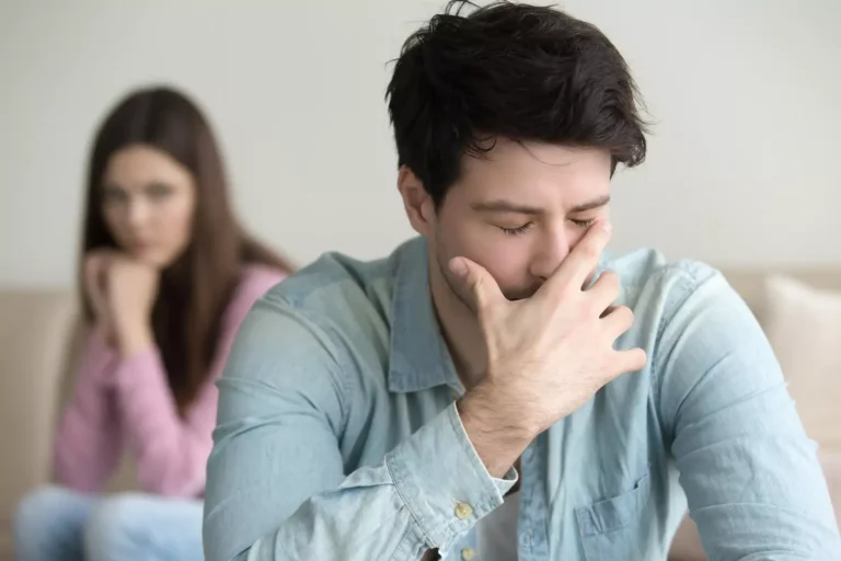 Photo of a young man emotionally hurt from an argument with his wife. The wife looks on from the background. Sometimes it's hard to tell if its a bad conflict or emotional abuse.