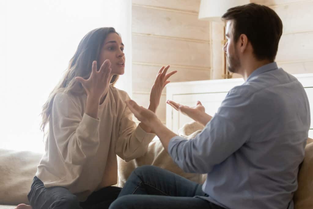 An angry young couple sit on a couch engaged in a heated argument.