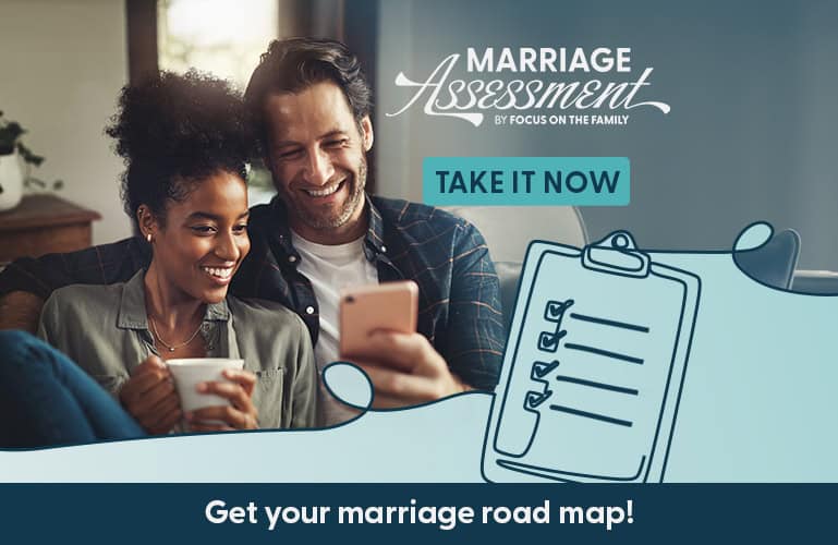 Smiling couple sitting together on a couch taking the Focus on the Family marriage assessment on a smartphone, with a coffee mug and marriage roadmap graphic in the image.