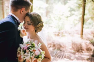 A newlywed couple embraces in a sunlit forest, with the groom gently kissing the bride’s forehead as she smiles with her eyes closed. The moment reflects love, intimacy, and the beauty of a new beginning, embodying heartfelt advice for newlyweds—to cherish and nurture their bond with tenderness and understanding.