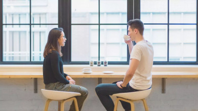 Photo of a woman initiating a conversation with her husband as they sit at a table in front of large windows while the man sips coffee and listens to her.