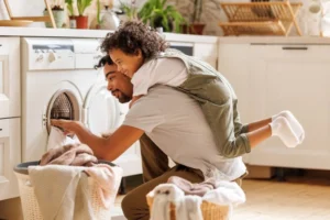 Side view of child in casual clothes with curly hair smiling and embracing dad loading washing machine during household routine in morning at home
