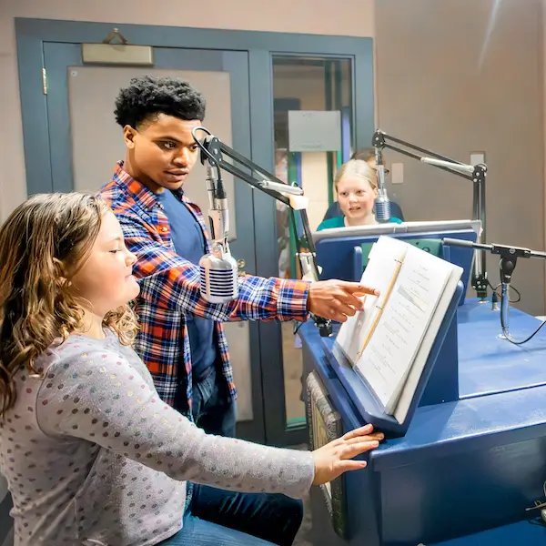 A young man helping two middle school girls record an audio play in a studio