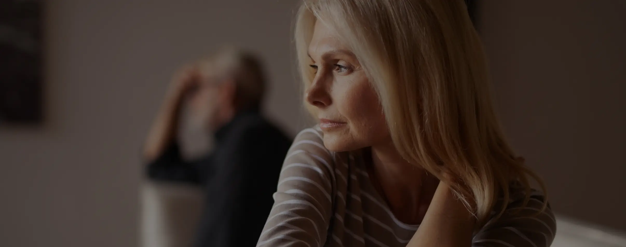 "A blonde woman looks contemplative in the foreground, while her husband appears faded in the background, symbolizing emotional distance in a struggling marriage."