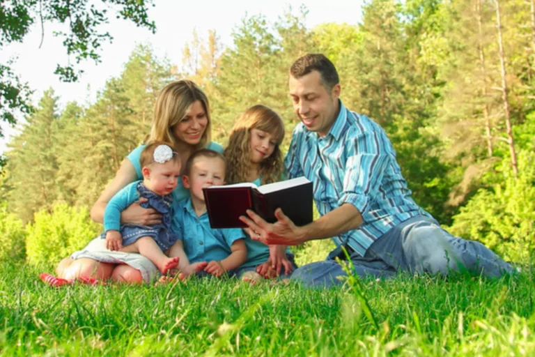 Public reading of scripture will help you grow spiritually as a family. Family gathered to gether under a tree reading the bible.