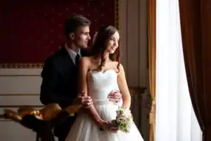Portrait of a newly married couple standing by a large window in an elegant room, the wife wearing a white wedding gown and holding a bouquet and the husband wearing a dark formal suit, as they look forward to their wedding night.