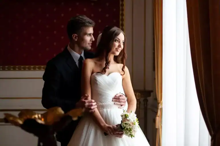 Portrait of a newly married couple standing by a large window in an elegant room, the wife wearing a white wedding gown and holding a bouquet and the husband wearing a dark formal suit, as they look forward to their wedding night.