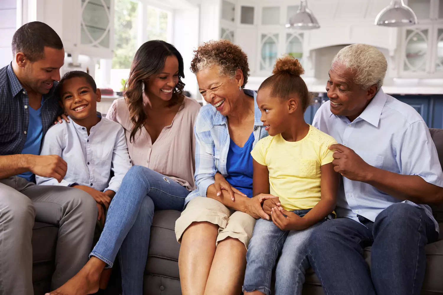 An extended African American family of two parents, two grandparents, and two kids sit on a couch enjoying each other's company. Setting boundaries with in-laws is important.
