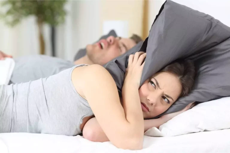 A frustrated woman lying in bed covers her ears with a pillow while her husband snores loudly beside her, symbolizing the struggle of dealing with your spouse’s character flaws.