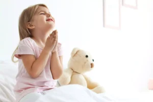 little girl with her teddy bear saying her bedtime prayers, sitting on her bed