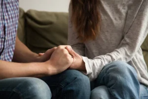 A couple sits next to each other on a couch, holding each other's hands tenderly in a marriage challenged by mental illness.