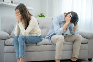 A distressed couple sitting apart on a couch, both with their heads in their hands, illustrating the challenges and need to learn how to handle stress in marriage.