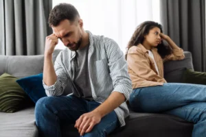 A couple sits on a couch, heads in hands, backs turned to each other, and clearly annoyed with each other. Spouses can become rivals in politically divided marriages.