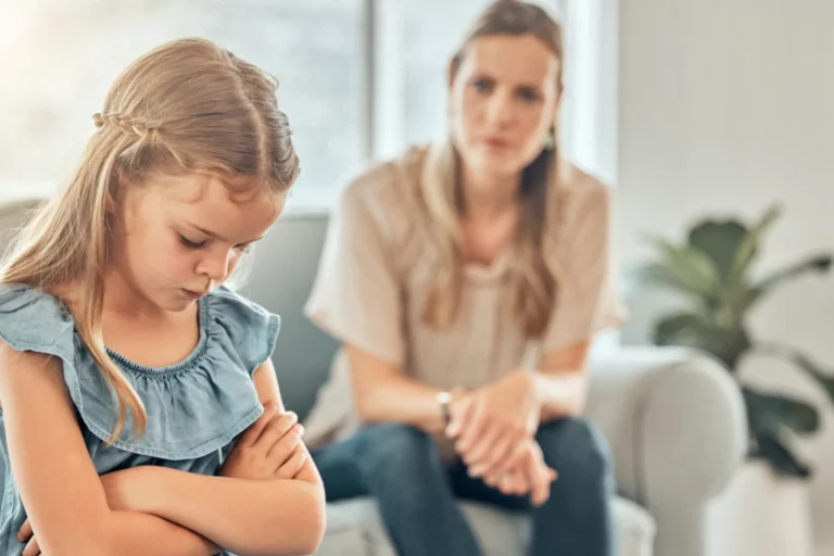 A young girl with her arms crossed, looking down, while an adult woman sits on a couch in the background, observing her. This scene represents disciplining kids.