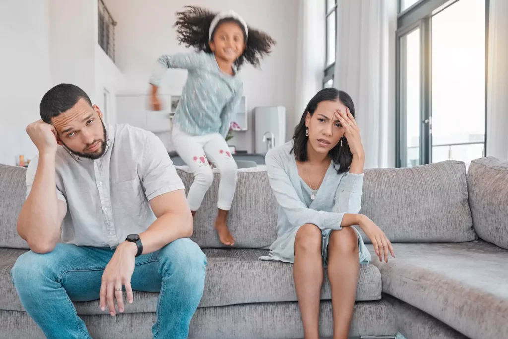 Photo of a man and woman sitting on a couch with their heads in their hands, clearly overwhelmed by their child, who is bouncing on the couch behind them. How you manage parenting stress and parental burnout in marriage can either harm your relationship or create deeper intimacy and happiness.