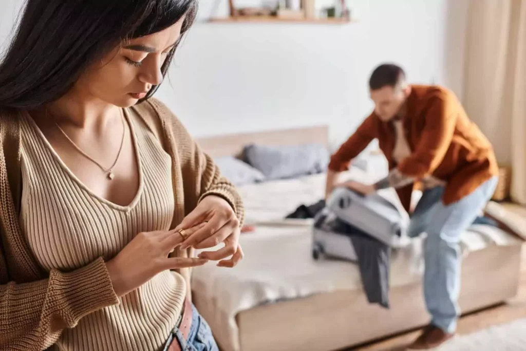 A woman stands in the foreground, looking down and twisting her wedding ring with a sorrowful expression, while in the background, her husband packs a suitcase on the bed. The image conveys emotional distress and uncertainty, representing the struggle of what to do if your spouse wants a divorce or separation.