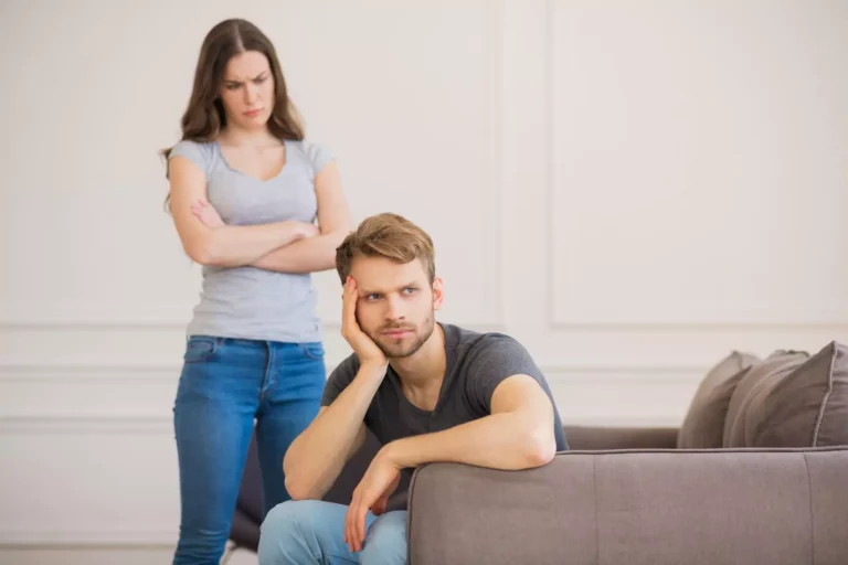 A woman stands, staring angrily at her husband. He sits on the couch, staring off into space in in a detached manner as they try to deal with autism in their marriage.