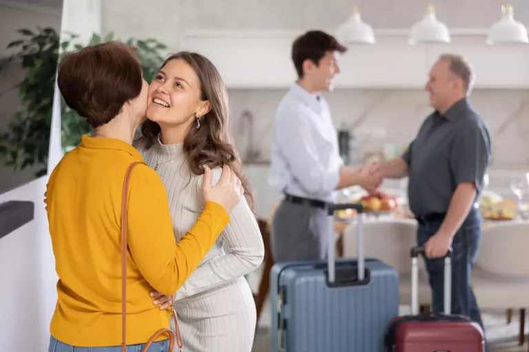 A young woman embraces her mother in the left foreground as her husband shakes hands with her father in the background. The husband has two suitcases, suggesting the woman is leaving home. A thriving marriage only works when both husband and wife leave home and cleave to each other in numerous ways.