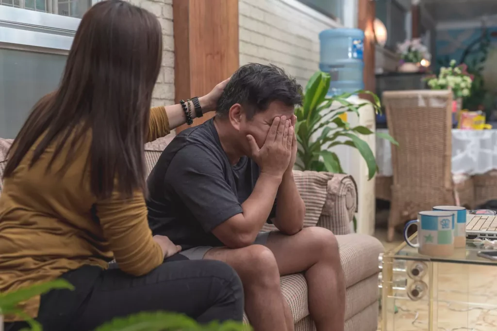 A man sits with his face buried in his hands, clearly sad. His wife sits next to him trying to comfort him. For victims of PTSD and their spouses, there is hope. Treatment and time can help mitigate PTSD's devastating impact on your marriage.