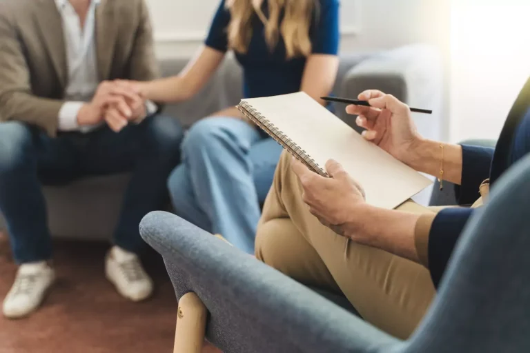 A photo of a couple in the background on a couch, holding hands lovingly. In the foreground, a professional marriage counselor takes notes on a notepad. The following considerations may help in locating a professional counselor and making the most of marriage counseling.