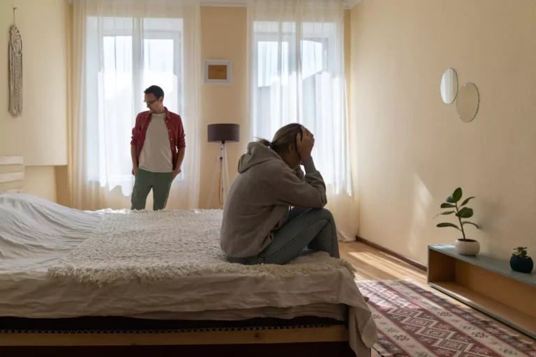 A man looks down at the floor in shame as his wife holds her head in her hands in grief and frustration as she sits on the edge of a bed. Pornography use is often symptom of a deeper issue, and using pornography is the way addicts avoid intimacy and cope with emotional pain.