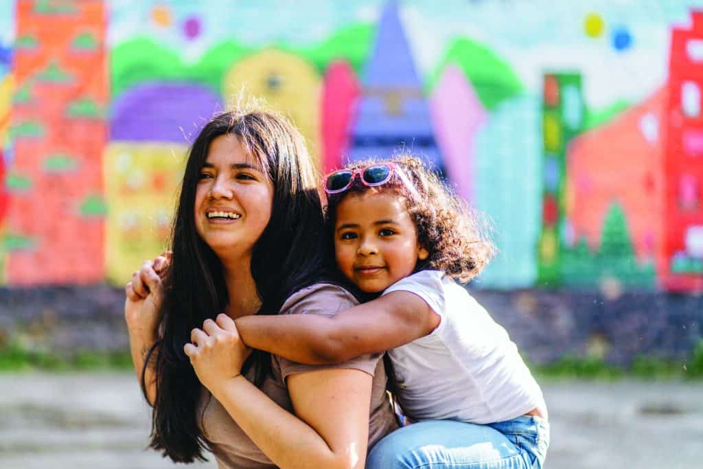 Enjoying ecuadorian mother playing and carrying young preschooler dark-skinned daughter on her shoulders while walking together in playground. Blurred children s drawing on background.