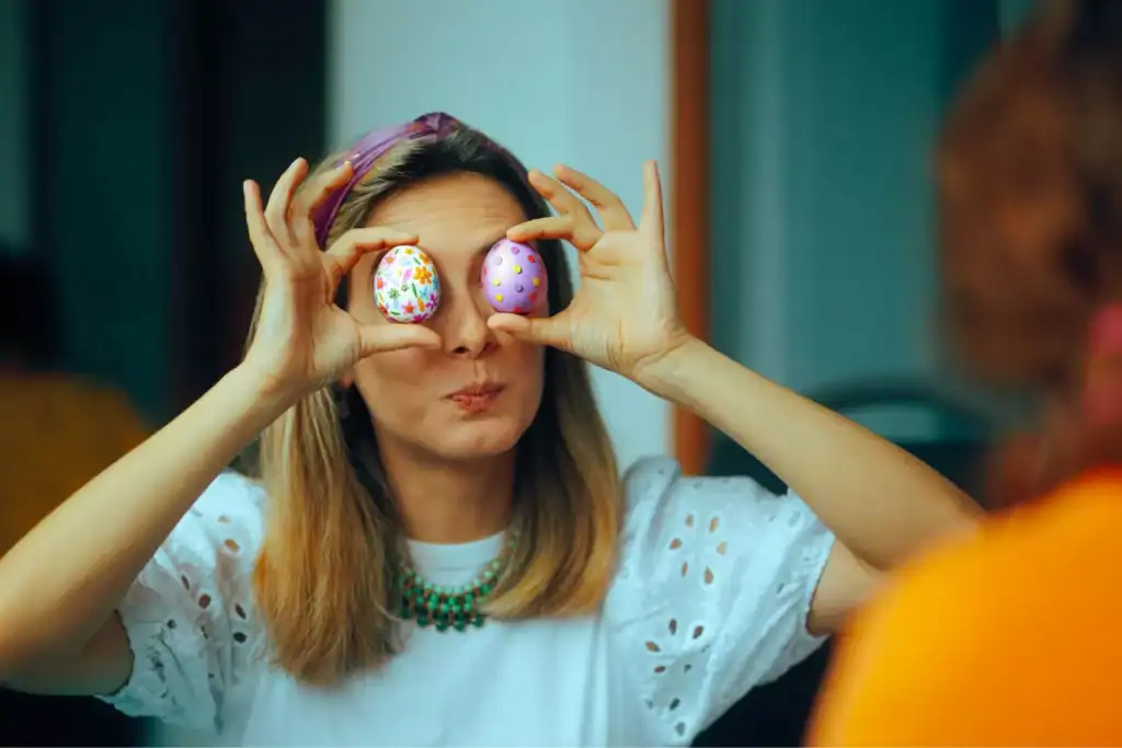 A woman wearing a white eyelet top holds two Easter eggs over her eyes, making a playful gesture during a conversation.