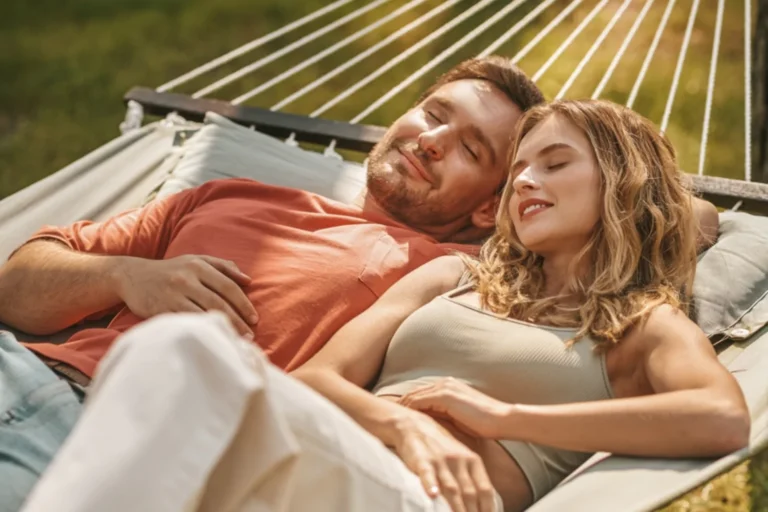 A smiling couple relaxes together on a hammock outdoors, enjoying a peaceful staycation moment.