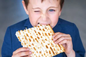 Young boy wearing a blue shirt, winking with one eye as he takes a big bite of a large piece of matzah bread.