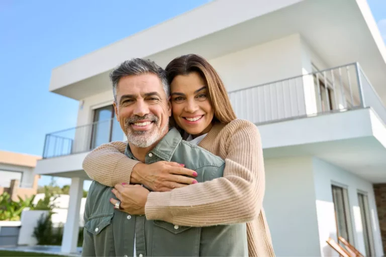 A happy couple embraces in front of their modern home, symbolizing building a strong marriage by giving grace and showing love.