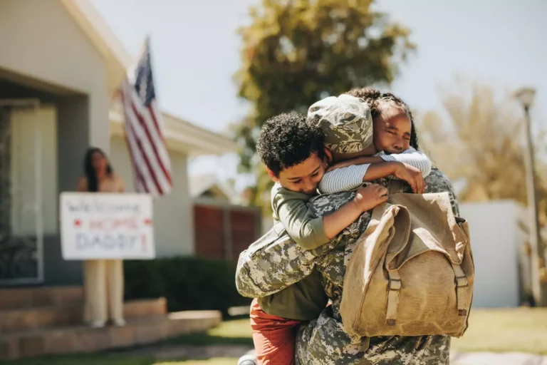 A heartwarming military homecoming as a soldier embraces two children tightly, with a welcoming sign and an American flag in the background.