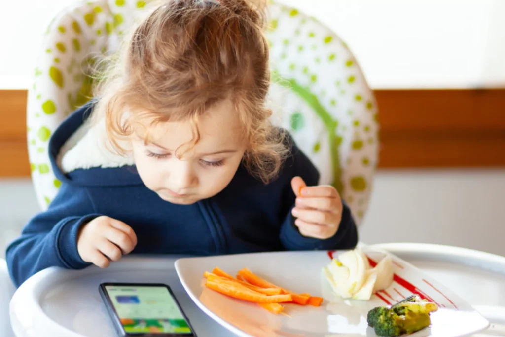 Young child sitting in a high chair, focused on a smartphone while eating carrot sticks and vegetables from a plate.