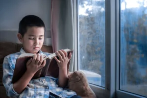 Young boy reading a Bible by the window, growing in faith through God’s Word.