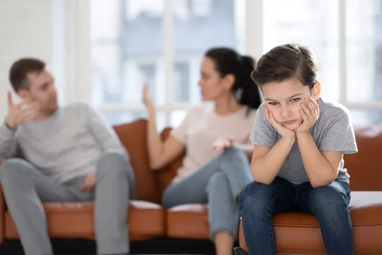 A young boy looks sad and frustrated while sitting apart from his parents, who are engaged in a tense discussion, illustrating challenges in disciplining kids in blended families.
