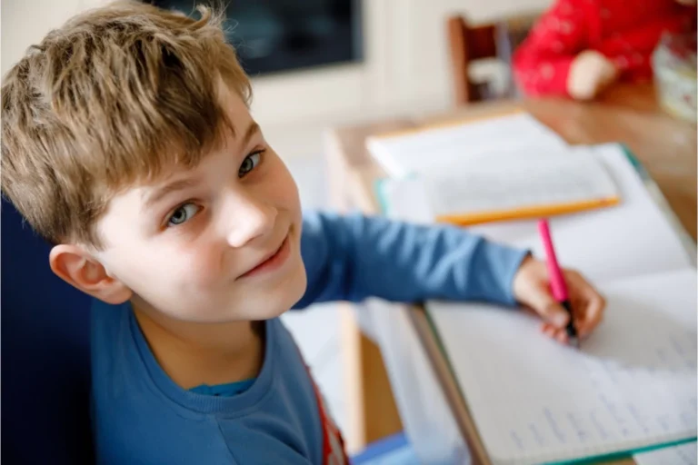 Smiling young boy homeschooling at kitchen table, writing in notebook
